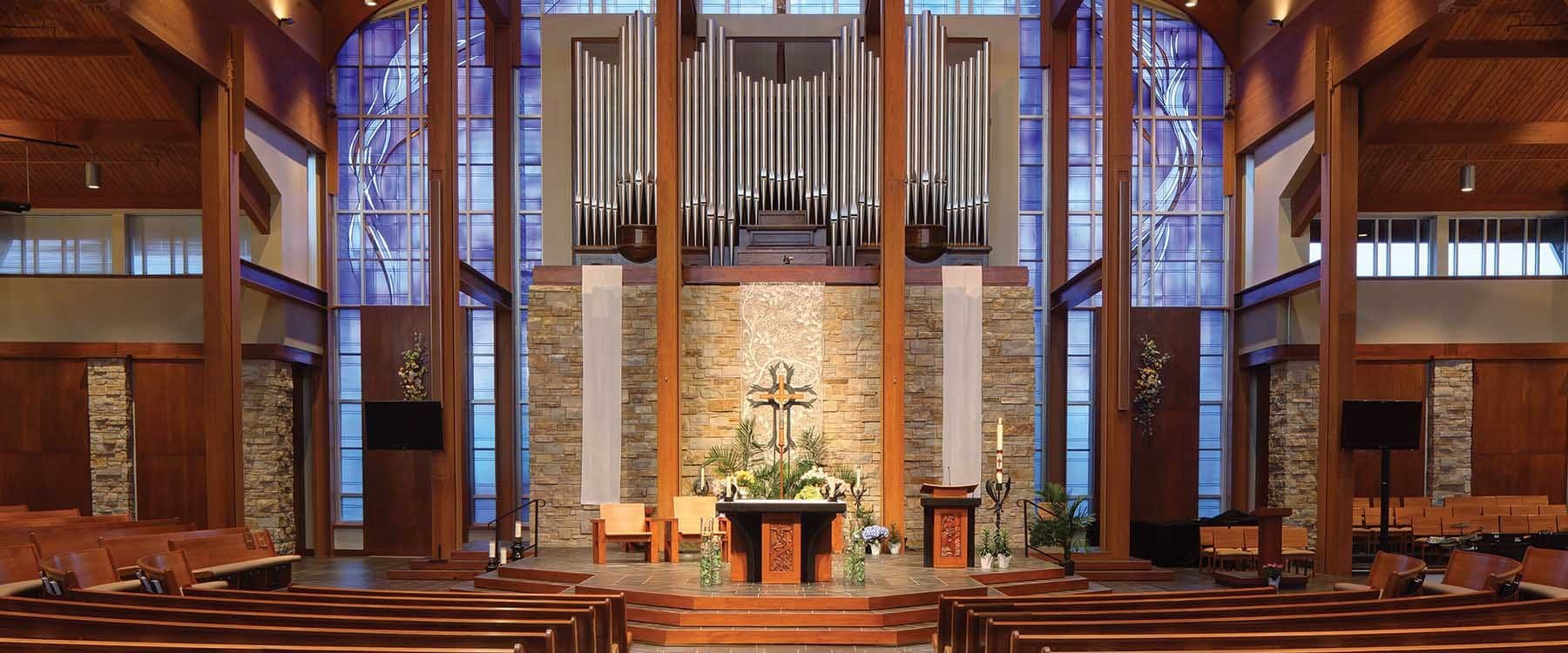 The Sanctuary for Holy Family Catholic Church in Fond Du Lac, Wisconsin combines natural materials include glue-lam beams, contemporary stained glass, simple Catholic Altar, and pipe organ pipes to provide an uplifting worship experience